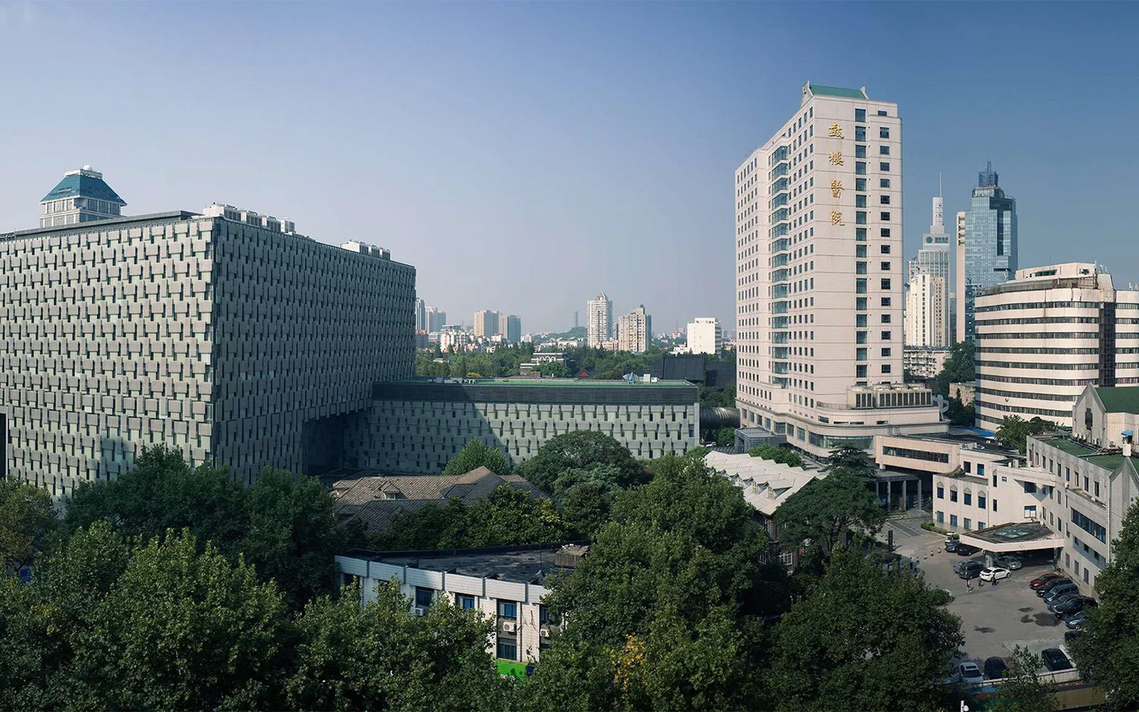 The Drum Tower hospital campus from above: a dark lattice-clad block on the left and a tower carrying the hospital's name in gold, with the city behind.